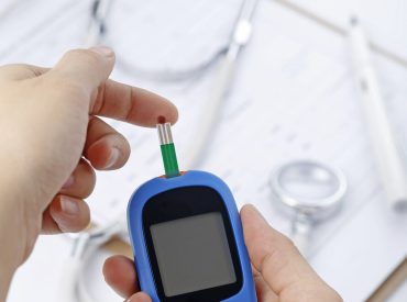 Hand holding a blood glucose meter measuring blood sugar, the background is a stethoscope and chart file Diabetes
